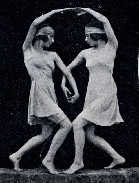 Black and white photograph. Two dancers pose with legs bent so their front knees touch. Each holds up one arm to make an arch. Their other arms curve and overlap, creating a heart-shaped form between their bodies.