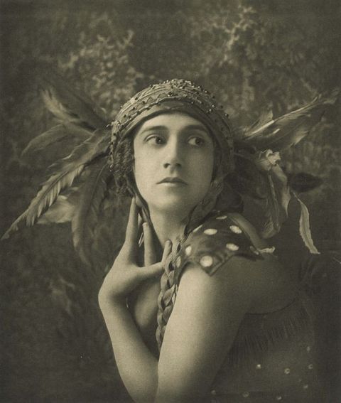 Black and white photograph. Shown from the waist up, a woman wearing a feathered headdress poses with her body facing our left as she turns her head to look off to our right. Both hands lie along her heck. The feathers fan out from the sides of her head like wings. Beaded silk wraps across her head like a turban. 