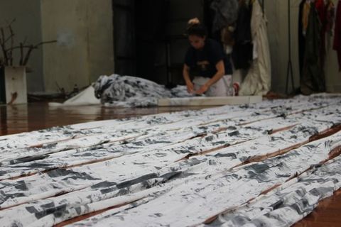 Colour photograph. About ten strips of white fabric streaked with dark grey lie along a wood floor. In the background and out of focus, a person kneels on the floor and works on more fabric piled there.