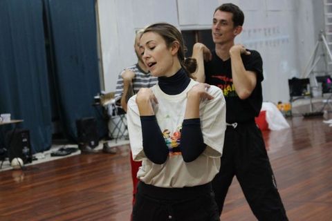 Colour photograph. Three people stand in a loose line coming towards us. They all bend their elbows to touch their shoulders, right hand to right shoulder and left to left. The woman at the front looks down and off to the side, and her mouth is open.