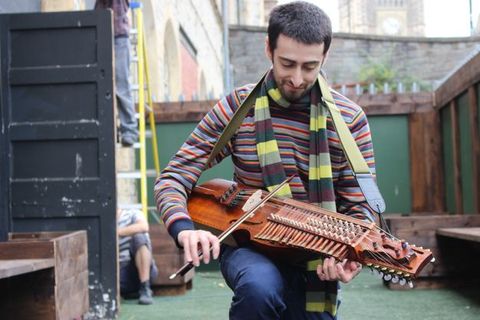 Colour photograph. A pale-skinned man with short black hair plays a nyckelharpa, which resembles a viola but has keys that change the pitch of the strings. The musician rests the instrument along his thigh, playing in a courtyard.